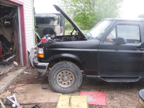 1996 Ford Bronco - A black 1996 Ford Bronco is parked in front of a garage with its hood up, while another vehicle with camouflage is visible behind it.
