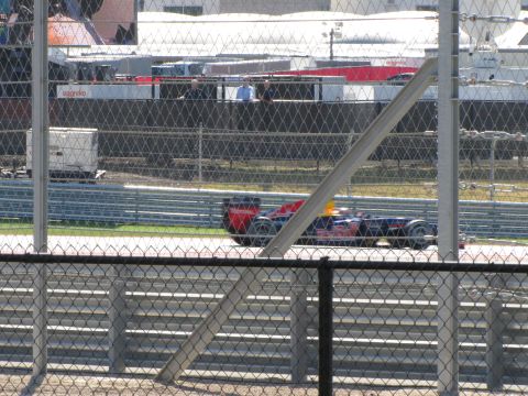 2006 BMW 5 Series - A Formula 1 race car speeds around a track, viewed through a chain-link fence.