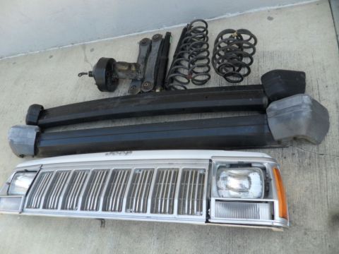 1994 Jeep Cherokee - Various parts from a Jeep Cherokee, including the grille, bumper sections, springs, and suspension components, are displayed on a concrete…