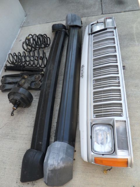 1994 Jeep Cherokee - Various Jeep parts including a grille, headlight, roof rails, and suspension components are laid out on a concrete surface.