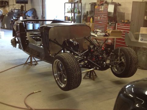 1975 Ford Ranchero - Three-quarter front view of an unpainted roadster hot-rod build on jack stands in a shop, with a bare aluminum firewall and body sides, no…