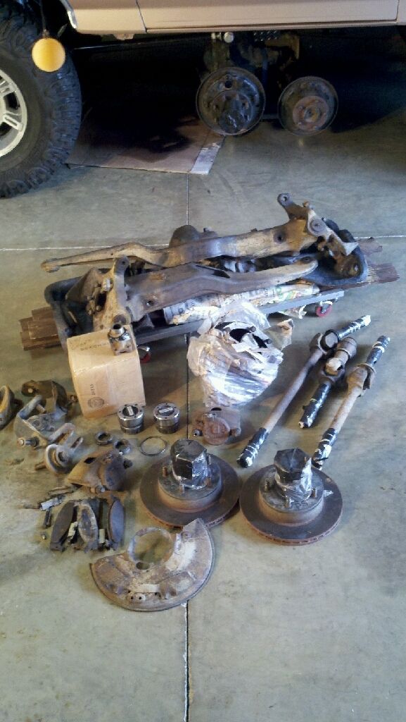 pile of rusty suspension and brake components, including control arms, brake rotors, and drive shafts, next to a vehicle's undercarriage