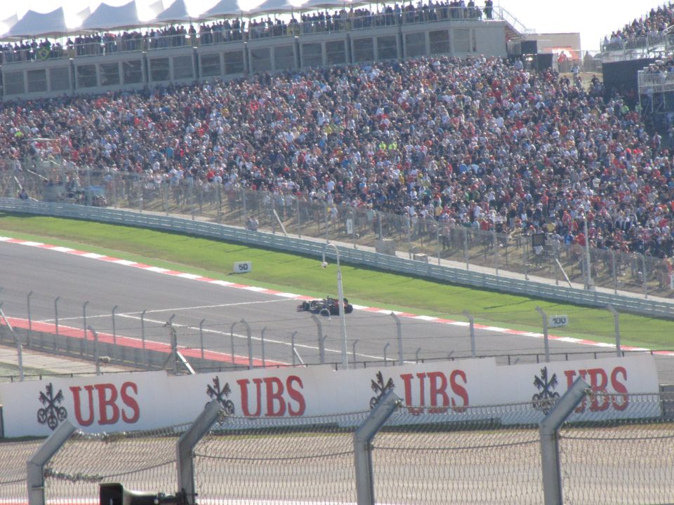 Race car on track with packed grandstands behind it. UBS logos visible on banners