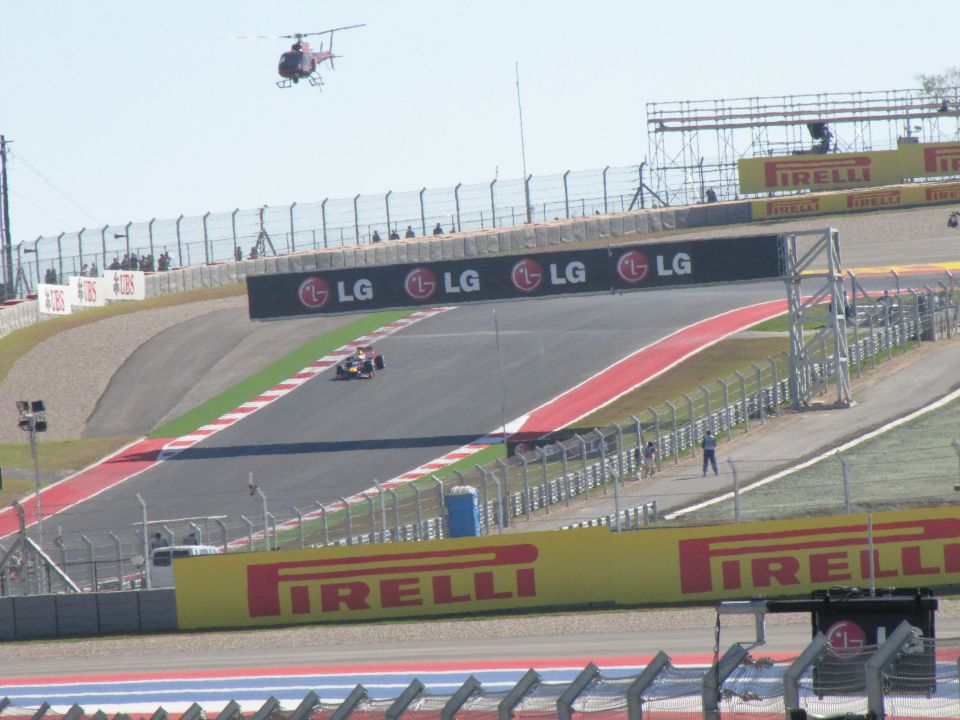Race track with a single-seater race car on the track and a helicopter in the sky. LG and Pirelli banners are visible