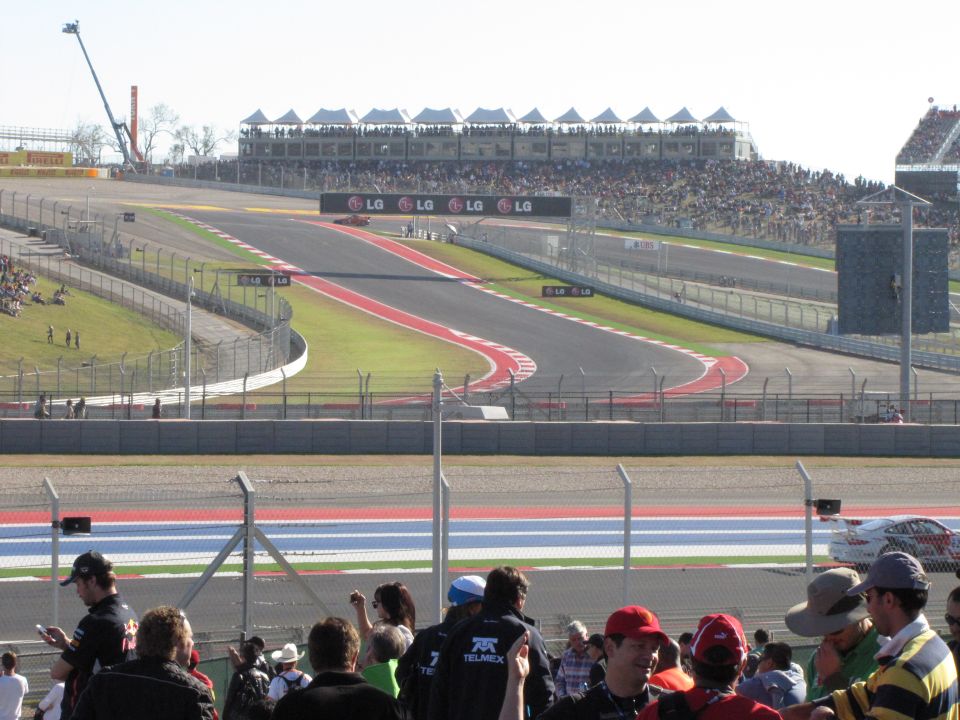 Race track with grandstands full of people. A few cars are on the track in the distance