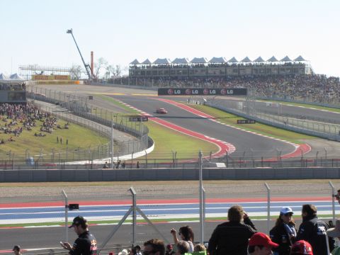 2006 BMW 5 Series - A lone red car navigates a sweeping turn on a race circuit, with crowds in the stands.