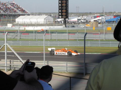 2006 BMW 5 Series - A race car speeds around the track, with spectators in the stands and others watching from the fence line.