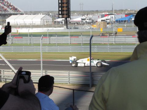 2006 BMW 5 Series - A white race car with green and blue accents speeds around a track, with spectators in the foreground and a scoreboard in the background.
