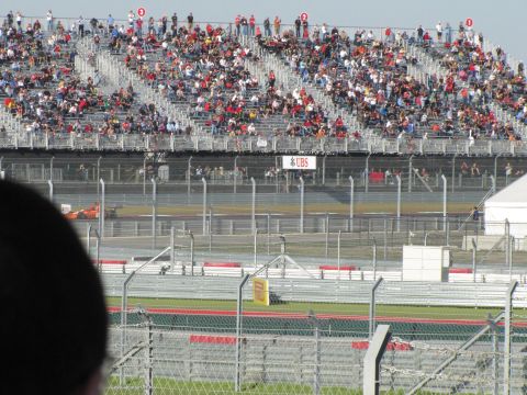 2006 BMW 5 Series - An orange race car speeds around a track, with a large crowd watching from the grandstands.