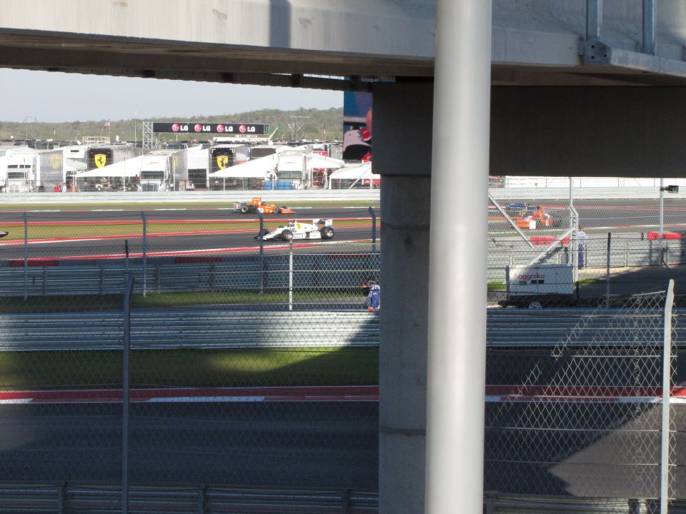 Two race cars on a track, one white and yellow, the other orange. Pit buildings in background