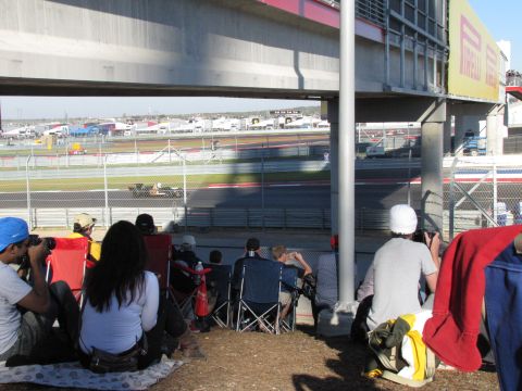 2006 BMW 5 Series - Spectators sit on the ground and in chairs watching a race car on a track, with a large structure overhead.