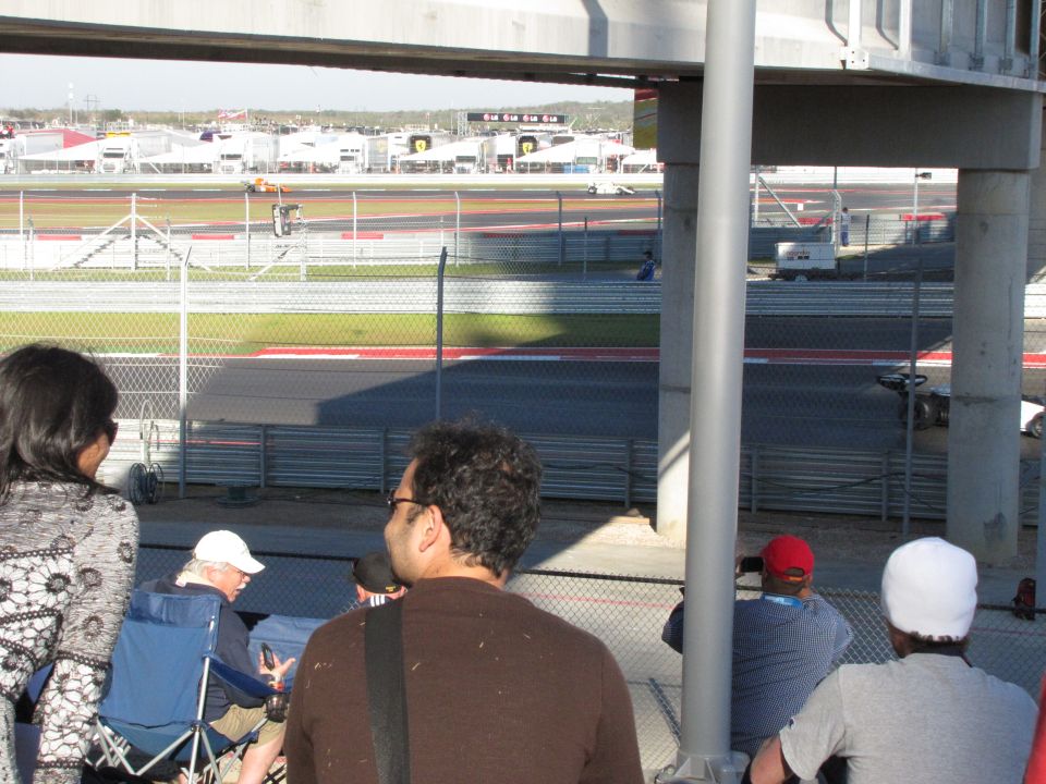 Spectators watch a race from a grandstand at a circuit, with buildings and a track visible in the distance