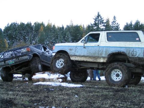 1988 Ford Bronco - Two heavily modified trucks tackle a muddy, snowy obstacle course in a wooded area.