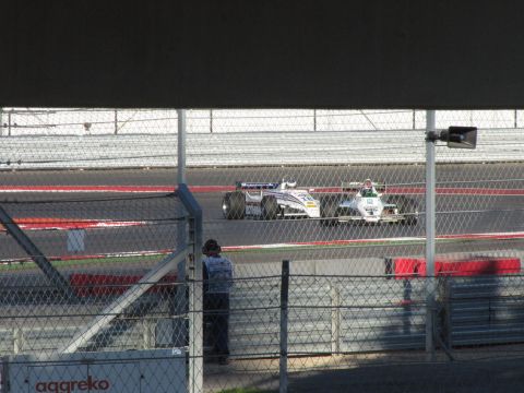 2006 BMW 5 Series - Two open-wheel race cars compete on a track, viewed through a fence.