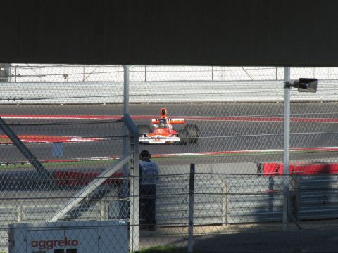 2006 BMW 5 Series - A Formula 1 car races on a track, viewed through a chain-link fence.