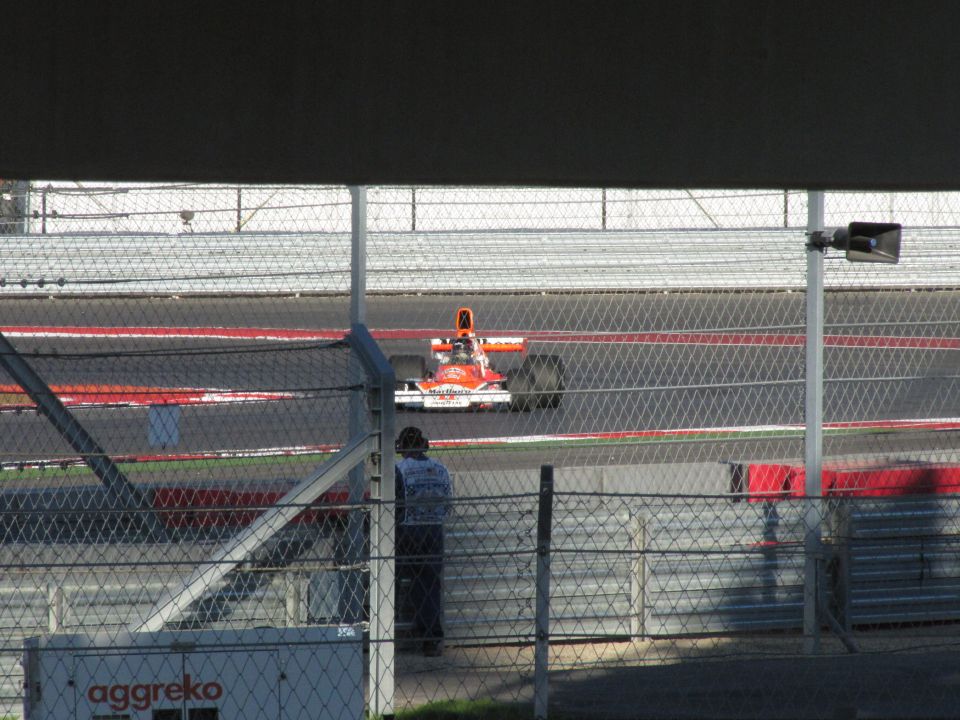 Orange Formula 1 car on a race track, seen from behind a fence. A person in a blue shirt stands near the fence