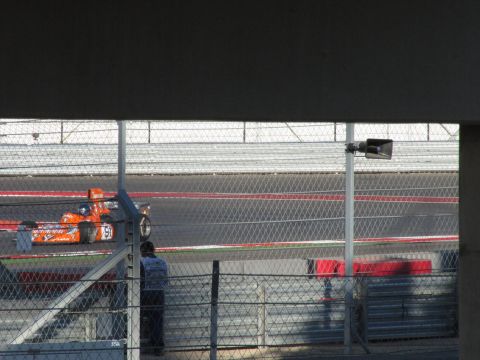 2006 BMW 5 Series - An orange race car with the number 91 is seen on a race track, with a spectator watching behind a fence.