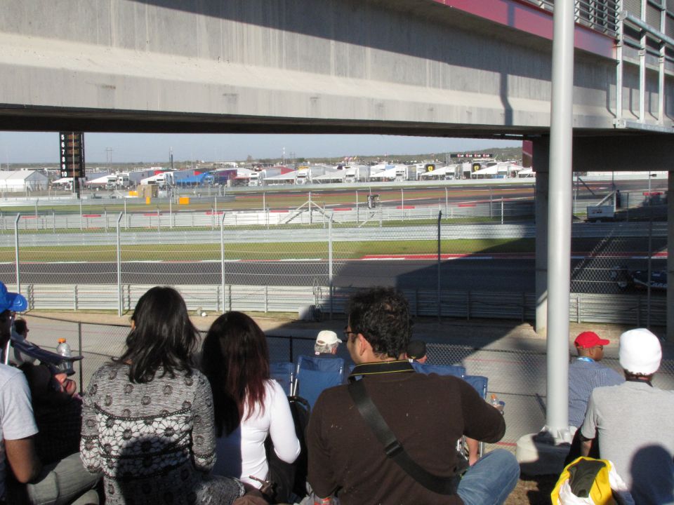 Spectators watch a race from the stands at a circuit, with buildings and track visible in the distance