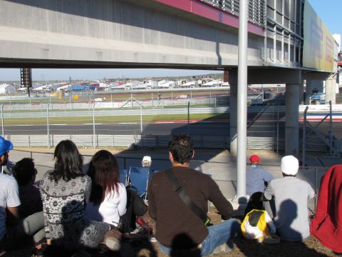 2006 BMW 5 Series - Spectators sit under a grandstand at a race track, with cars visible on the circuit in the distance.
