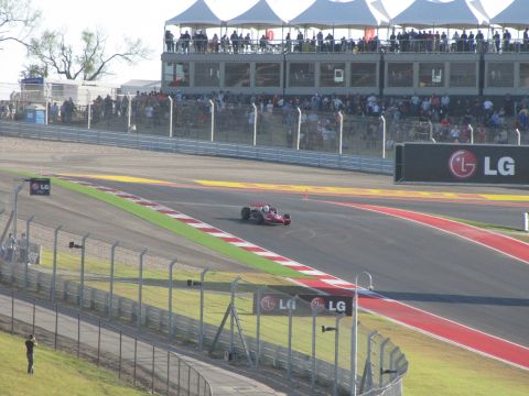 2006 BMW 5 Series - A single-seater race car navigates a turn on a sunny day at a racetrack with grandstands full of spectators.