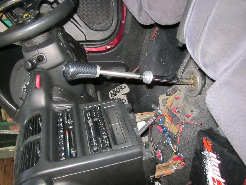 Car interior showing a carbon fiber-wrapped gear shift knob on an extended shaft, with exposed wiring and pedals visible