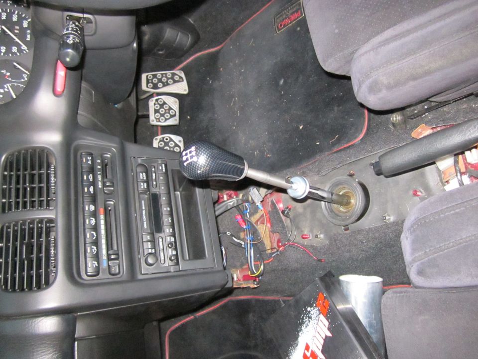 Car interior showing a carbon fiber shift knob on an extended shaft, with aftermarket pedals and exposed wiring