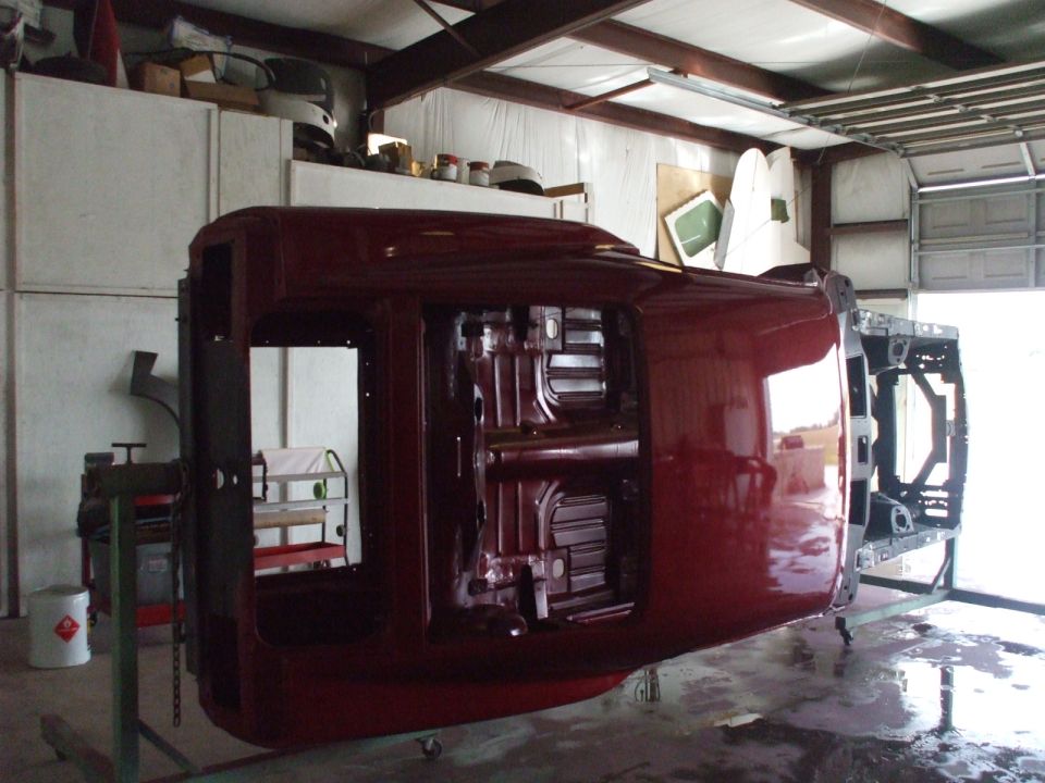 Maroon car body upside down on a metal frame, showing the underside and interior, in a workshop