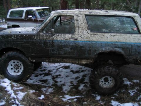 1988 Ford Bronco - A muddy Ford Bronco is seen off-road with another vehicle in the background.