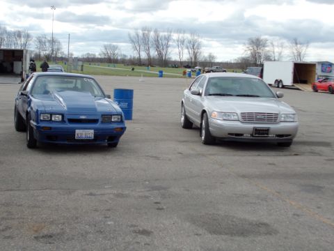 2003 Ford Crown Victoria - A silver Ford Crown Victoria and a blue Ford Mustang drag car are parked side-by-side at a drag strip.