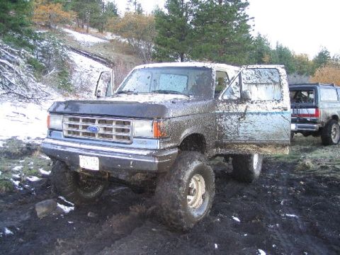 1988 Ford Bronco - Two Ford Broncos are stuck in the mud with snow on the ground and trees in the background.