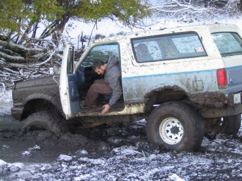 1988 Ford Bronco - A white and blue SUV is stuck deep in mud, with a person exiting the driver's side door.
