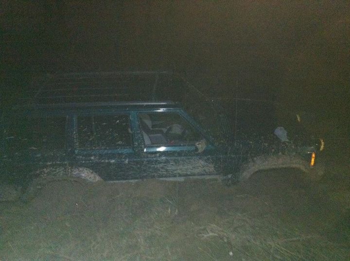 Mud-covered green Jeep Cherokee stuck in dirt, side view, dark conditions