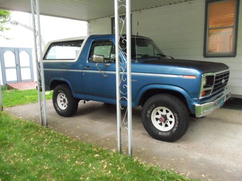 1985 Ford Bronco - A blue Ford Bronco with a white top is parked under a carport, showing its front quarter profile.