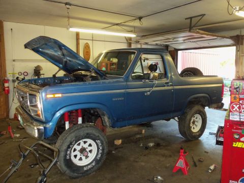 1985 Ford Bronco - A blue 1985 Ford Bronco is shown in a garage with its hood open and front end elevated on jacks, suggesting maintenance or repair work.