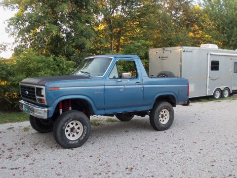 1985 Ford Bronco - A blue Ford Bronco is parked on a gravel surface, hitched to a silver enclosed trailer.