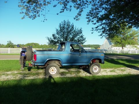 1985 Ford Bronco - A blue 1985 Ford Bronco is parked on a grassy area next to a road, with a dirt bike mounted on a carrier at the rear.