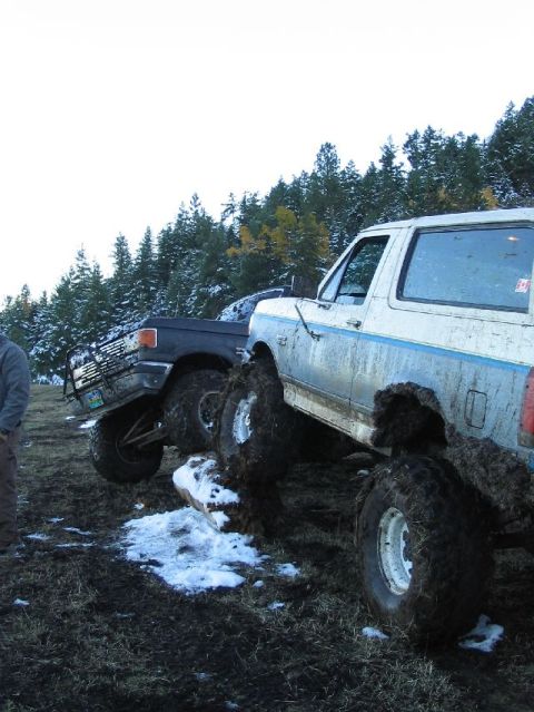 1988 Ford Bronco - Two off-road vehicles are stuck in thick mud and snow, with trees in the background.