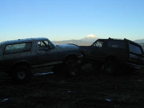 1988 Ford Bronco - Two Ford Broncos are parked on a muddy incline with a snow-covered mountain visible in the distance under a clear sky.