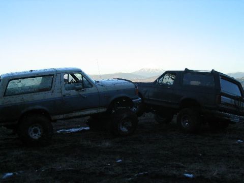 1988 Ford Bronco - Two modified Ford Broncos are positioned on a muddy terrain under a clear sky with distant mountains visible.