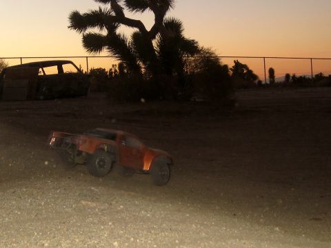 1996 Ford Bronco - An orange RC truck navigates a dirt path as the sun sets, with a derelict van visible in the background.