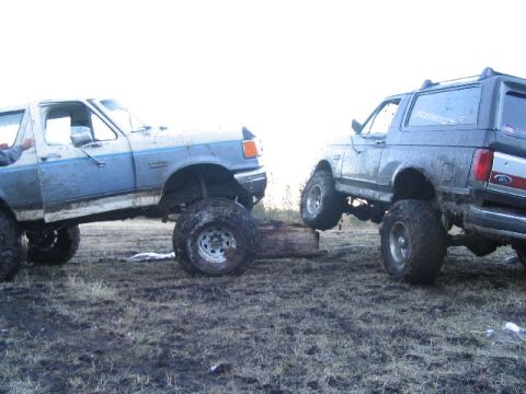 1988 Ford Bronco - Two Ford Broncos, one blue and white and the other gray, are shown navigating a muddy off-road course.