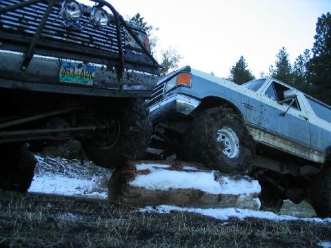 1988 Ford Bronco - Two heavily modified SUVs are navigating an obstacle course, with one vehicle perched on top of a fallen log.