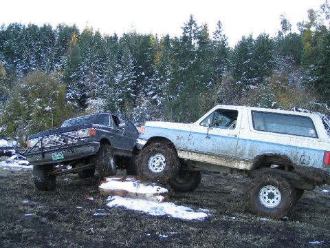 1988 Ford Bronco - Two heavily modified SUVs are stuck in a muddy, snowy terrain, with one vehicle attempting to drive over a log.