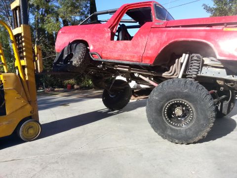 1978 Ford Bronco - Forklift lifting a modified red 1978 Ford Bronco with custom suspension and large off-road tires.