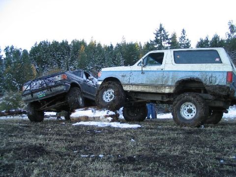 1988 Ford Bronco - Two heavily modified trucks navigate challenging terrain in a snowy, wooded area.