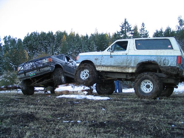 Two lifted trucks, one a blue and white Bronco, the other a dark pickup, are off-roading in a muddy, snowy field with trees in the background