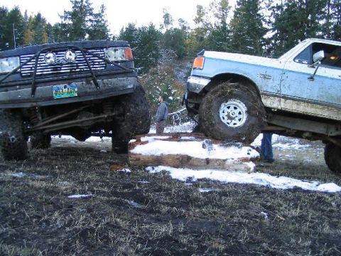 1988 Ford Bronco - Two heavily modified trucks are navigating an obstacle course in a muddy, wooded area.