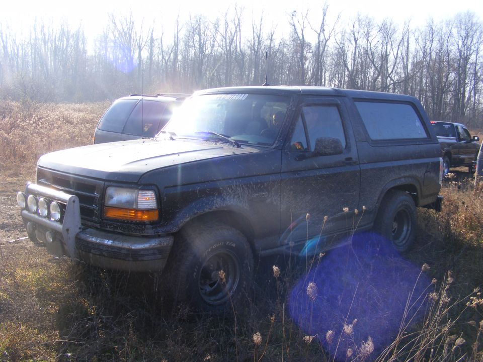 Dark Ford Bronco with brush guard and auxiliary lights parked in a field with trees in the background. Another SUV and truck are visible
