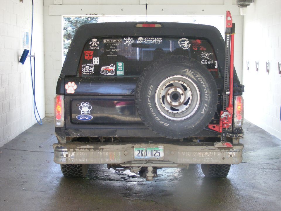 Rear view of a black Ford Bronco with a spare tire mounted on the back, covered in stickers, and a high-lift jack on the side