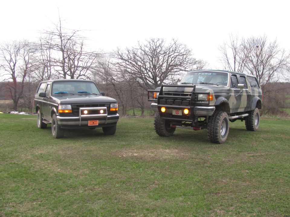 Two Ford Broncos parked on grass, one black, one camo with a bull bar